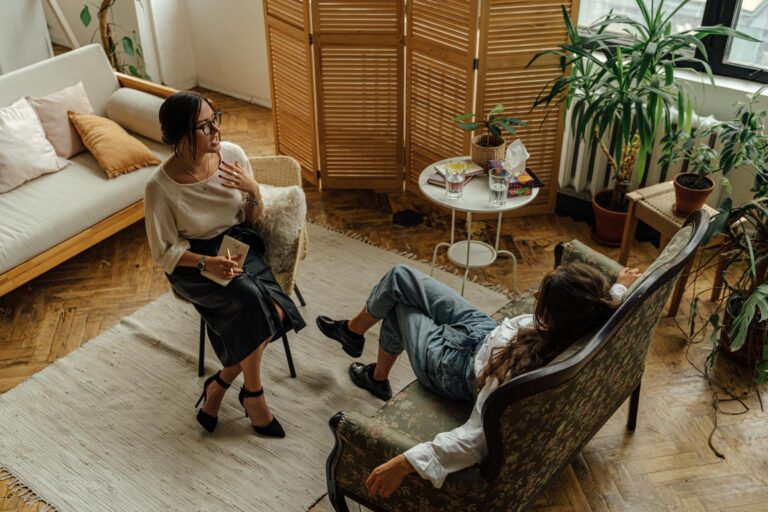 Two women engaged in a psychotherapy session in a warm, inviting interior with plants and natural lighting.